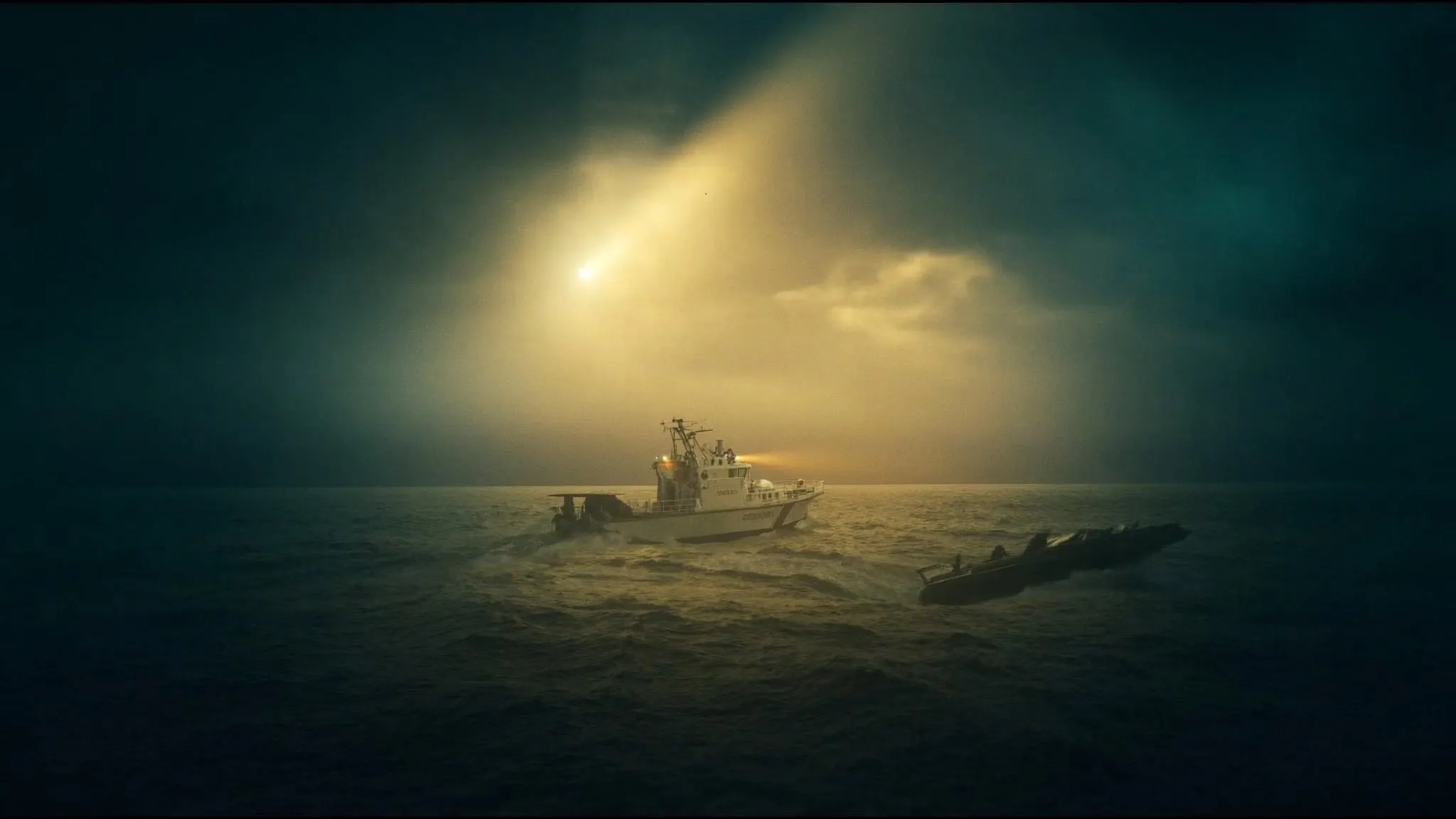 A small boat running dark at night under a searchlight from a pursuing patrol.